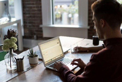 A man sat at a brown table on his laptop, doing some work.