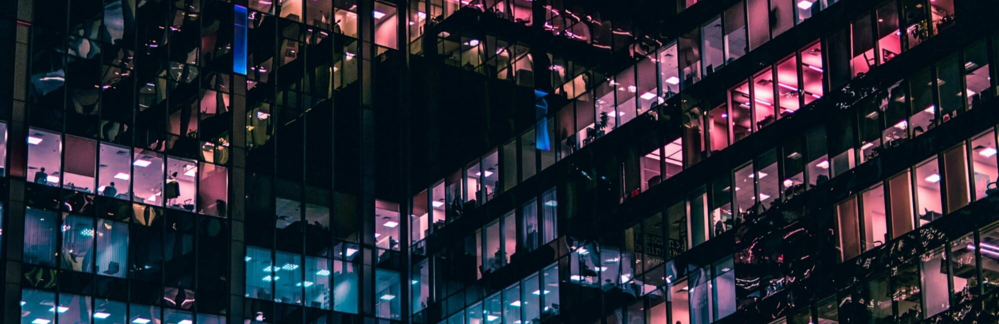 A multi-story glass office building at night with illuminated windows displaying various colors, including pink and blue, creating a geometric pattern of reflections.