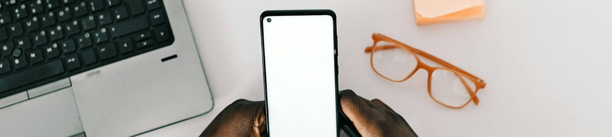 Smartphone with blank screen in hands of African American man at white desk, with phone, laptop, and glasses nearby.